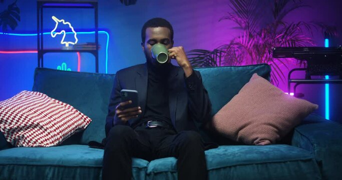Attractive young African American stylish man tapping on smartphone while drinnking hot tea on couch in living room with neon lights. Handsome black guy sending messages on his phone while chatting