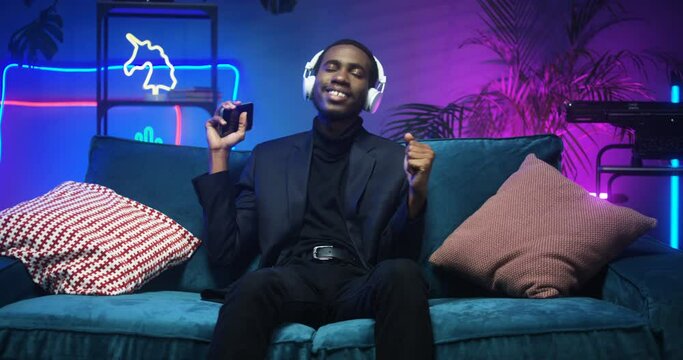 Attractive young African American stylish man in headphones listening to music on smartphone player while sitting on couch in futuristic living room. Black guy enjoying song and dancing on sofa in