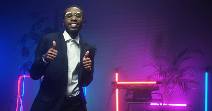 African American young handsome man in glasses and fancy suit dancing in room with neon lights. Black good looking gentleman at futuristic party. Guy in blue and pink lights of asteras. Male funny