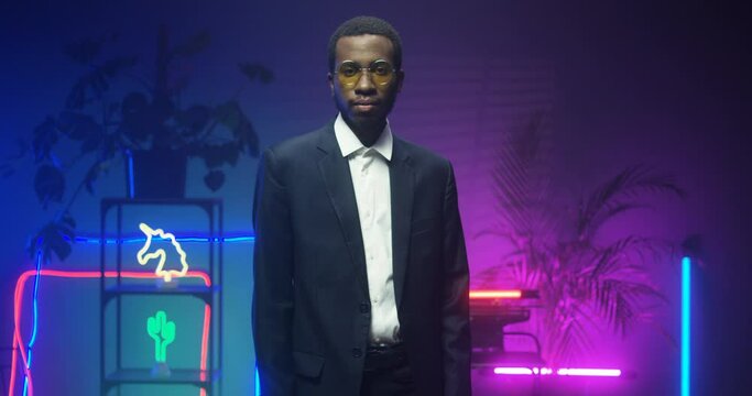 Portrait shot of African American serious handsome man in glasses and suit looking straight at camera in futuristic room with blue and pink lights of neon tubes. Young guy with strict face in club