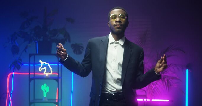 African American young handsome man in glasses and fancy suit dancing in room with neon lights. Black good looking gentleman at futuristic party. Guy in blue and pink lights of asteras. Male funny