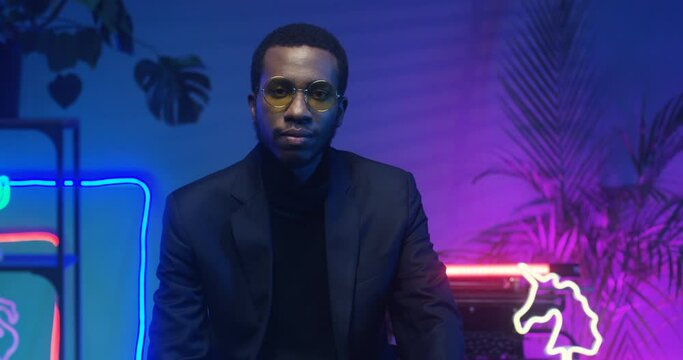 Portrait of African American serious handsome man in glasses looking straight at camera while sitting in futuristic room with blue and pink lights of neon tubes. Young guy with strict face in club