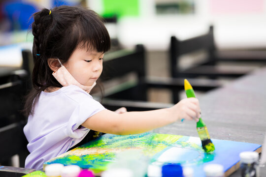 Cute Asian girl is coloring on a canvas. Child grab a large paintbrush, paint and make art in classroom. Happy little girl open the cloth mask from his mouth and nose. Kid are 3 years old.