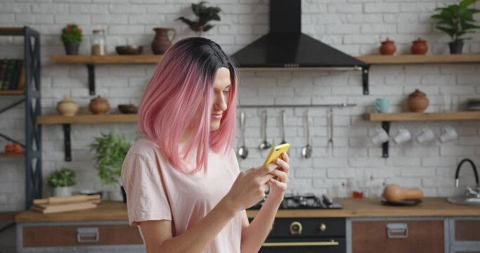 Cheerful young trans-woman with stylish pink wig surfs internet with contemporary cellphone standing in decorated kitchen closeup