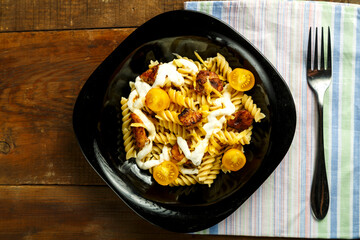 Pasta with chicken and cheese in a black plate on a wooden table on a napkin with a fork.