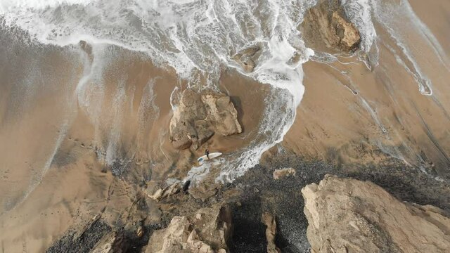 surfer with surfboard walking on sand along the beach and ocean waves and leaving footprints in the sand. Aerial drone cinematic shot.