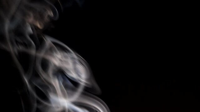 Smoke streaming in the dark from a burning incense stick. Whimsical curls of smoke on a dark background. A soothing atmosphere of relaxation.