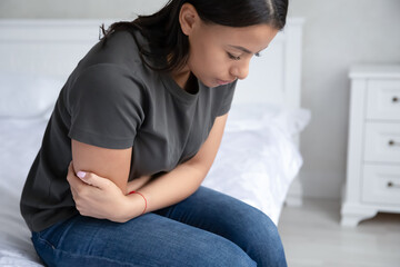 Sick young African American woman sit on bed at home hold belly suffer from abdominal pain or stomachache. Unhealthy biracial female struggle with painful ache, have intoxication or poisoning.
