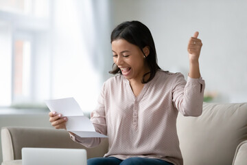 Excited young biracial woman sit at couch at home triumph reading good news in paper letter or...