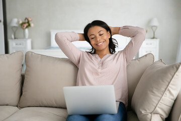 Happy young biracial woman sit relax on couch in living room watch video on laptop gadget online. Smiling African American female tenant renter rest on sofa at home using computer, enjoy weekend.
