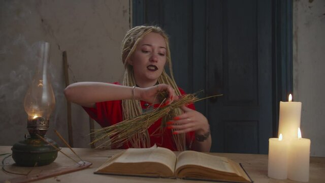 Cruel attractive female voodoo practitioner with dreadloks casting black spells, imposing curse and breaking voodoo doll, causing harm and damage to person while conjuring in rural house.
