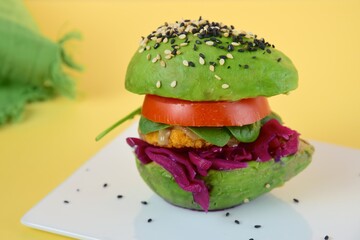 Vegan avocado burger with chickpea patty, tahini, spinach, red cabbage, tomato and sesame seeds on yellow background