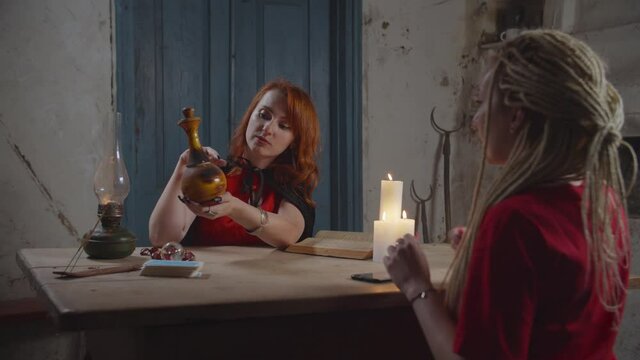 Pretty mysterious white witch in black cape giving rustic vial with love potion to hopeless woman in love, to attract beloved man during seance of fortune telling in dimmed rural house.