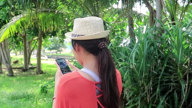 Young girl in hipster style cloths & sun hat using mobile phone or smartphone at rainforest green garden park by ocean sea beach in tropical summer or spring sunlight at sunshine day, B-roll footage
