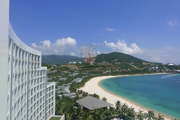 Fototapeta premium Top view of the turquoise sea, the curved line of the sandy beach, mountains. A fragment of a tall building, several villas, is visible. In the distance a colorful Ferris wheel. Vietnam. Nha Trang.