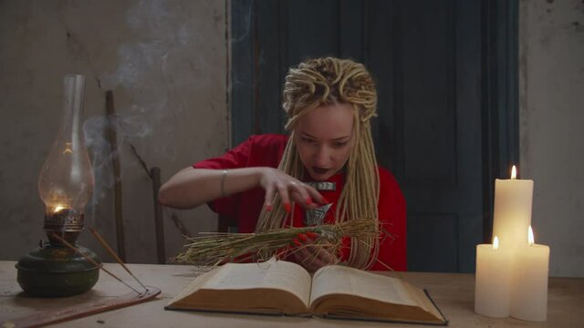 Evil eerie voodoo sorceress with dreadlocks practicing black magic , casting curse on straw voodoo doll, furiously piercing voodoo doll with fork, causing harm and damage to person as act of revenge.