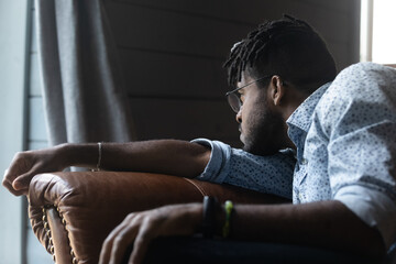 Upset young African American male sit on sofa at home look in distance mourning missing. Unhappy sad biracial guy distressed with life problems, feel lonely thinking or pondering. Solitude concept.