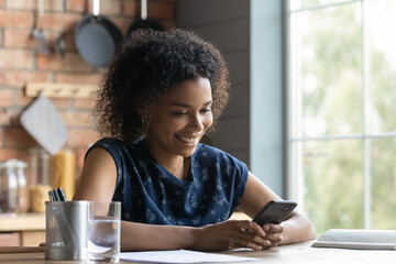 Smiling African American woman sit at table at home office use modern cellphone gadget texting or messaging. Happy biracial female user browse wireless internet on smartphone. Communication concept.