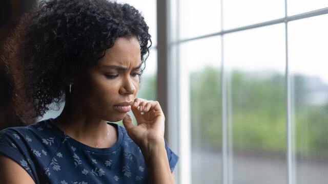 Close up pf unhappy 20s biracial woman stand near window miss or mourn at home. Sad depressed young African American female feel loneliness solitude, lack communication. Loner, outcast concept.
