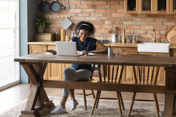 Young African American female student in headphones sit at home kitchen study online on laptop. Millennial biracial woman take distant course or training, watch webinar on computer, make notes.