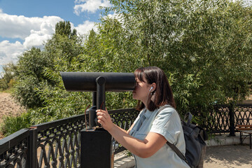 Close-up portrait of a young woman in light clothing on a clear, warm day in a city park. Looks through binoculars at the observation deck. Weekend rest. Selective focus.