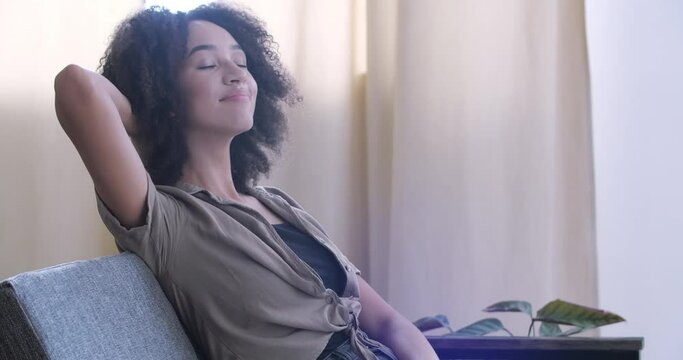 African american ethnic woman rest on couch in living room puts hands behind head leaned on sofa opens eyes looking at camera feels good. Lazy weekend day, enjoying cozy home atmosphere concept