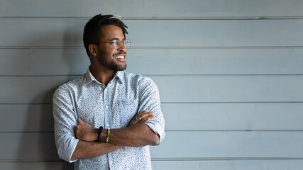 Happy 20s African American man in spectacles isolated on grey wooden wall background look in distance dreaming thinking. Smiling young biracial male planning or visualizing at home. Vision concept.