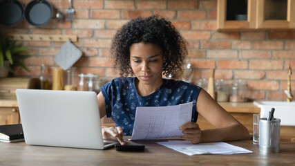 Serious millennial biracial woman calculate household expenditures finances on calculator at home. Focused African American wife pay bills taxes online on laptop, manage family finances or budget. - obrazy, fototapety, plakaty