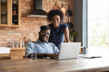 Portrait of smiling young African American family renters sit at table at modern design home kitchen using laptop. Happy ethnic biracial couple tenants pay household bills taxes on computer online.