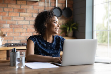 Smiling young biracial woman work on laptop at home office look in distance dreaming or visualizing. Happy African American female use computer feel distracted thinking or planning. Vision concept.