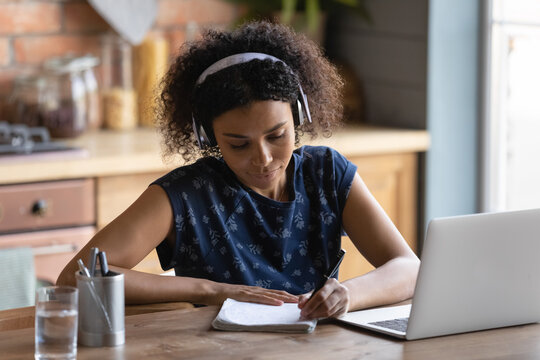 Smart millennial biracial girl student in earphones study distant on laptop from home make notes. Young African American woman watch webinar, take web course on computer. Online education concept.