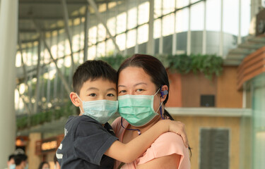 A happy mother is holding the young kid and looking at the camera. Both parent and child are wearing the disposal/surgical mask and standing indoor. An Asian mom and boy are hugging together with love