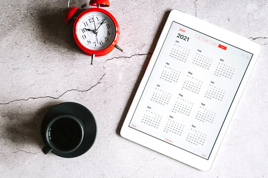a tablet with an open calendar for 2021 year, a cup of coffee,  and a red alarm clock on a gray concrete background