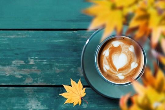 Drinking Coffee in Fall and Autumn Season. Hot Coffee Latte Cup on Blue Wooden Table. Top View. Focus on Latte Art in Heart Shape. blurred Yellow Maple Leaf as foreground