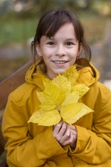 Deurstickers Leuk meisje in gele jas zittend op de bank. Het meisje houdt een armvol bladeren vast © Albert Ziganshin
