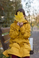 Foto op Aluminium Leuk meisje in gele jas zittend op de bank. Het meisje houdt een armvol bladeren vast © Albert Ziganshin