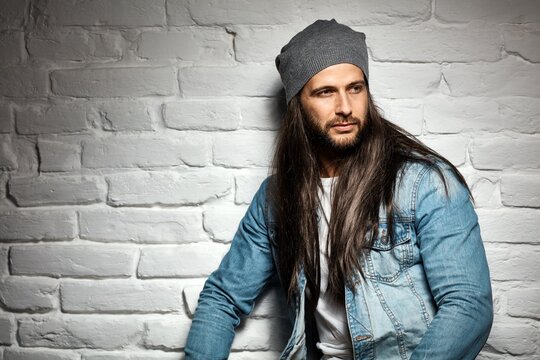 Portrait Photo Of Rocker Guy In Sunglasses And Woolly Hat Standing Against Brick Wall. 