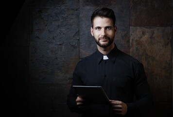Portrait of handsome young catholic priest using tablet against dark background.