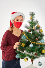 Young brunette Spanish woman dressed in Santa hat celebrating Christmas with gifts and decorating the Christmas tree, covid-19, with a red facing mask