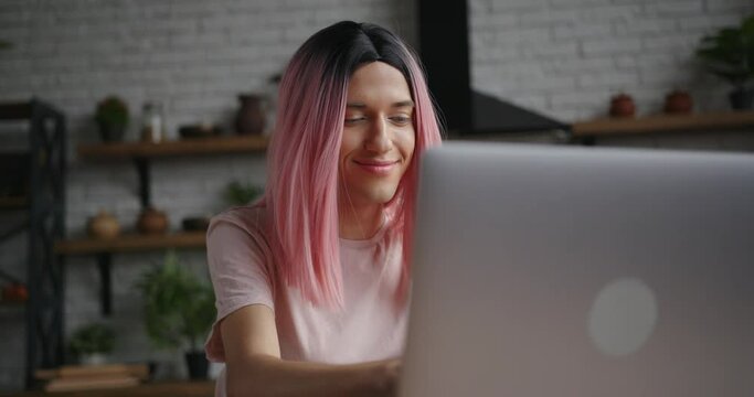 Attractive young transwoman with pink wig types message on laptop and smiles sitting at table in decorated kitchen at home