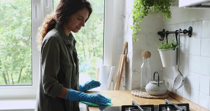 Housewife keeps home in tidy, wear rubber gloves use rag cleans countertop standing in cozy domestic kitchen do chores smiles enjoy routine process. Housekeeping services agency worker working concept