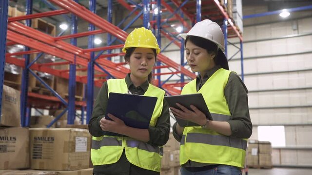 woman manager in white hard hat talking, showing tablet and training new girl employee in the factory warehouse