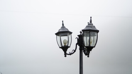 Fototapeta na wymiar Top of a black street lamp and gray fog around, place for text. The concept of a cold wet summer morning