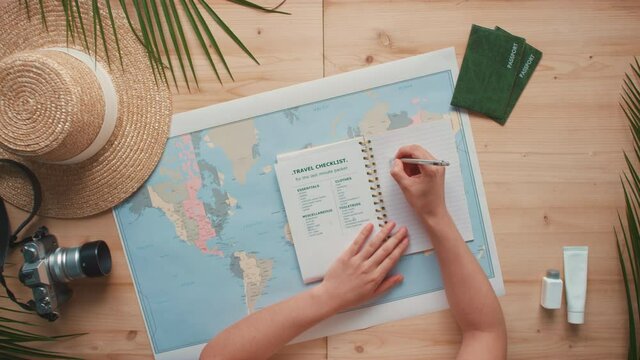 Top-view horizontal flat lay of female hands keeping bullet journal with travel checklist inside