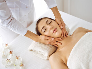 Obraz na płótnie Canvas Beautiful brunette woman enjoying facial massage with closed eyes. Relaxing treatment in medicine and spa center concepts