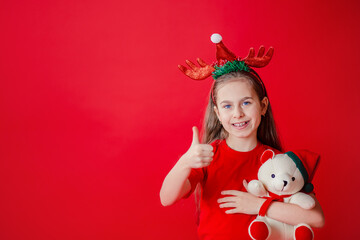 Portrait of a funny cheerful girl with a bandage of horns on her head hugging a teddy bear in Christmas pajamas isolated on a bright red background.