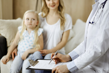 Doctor and patient. Pediatrician using clipboard while examining little girl with her mother at home. Sick and unhappy child at medical exam. Medicine concept