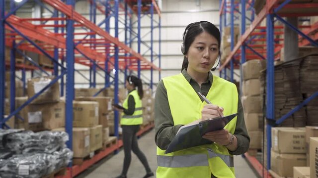 two asian chinese women workers wearing headset and talking in distribution on online phone call in large stockroom.