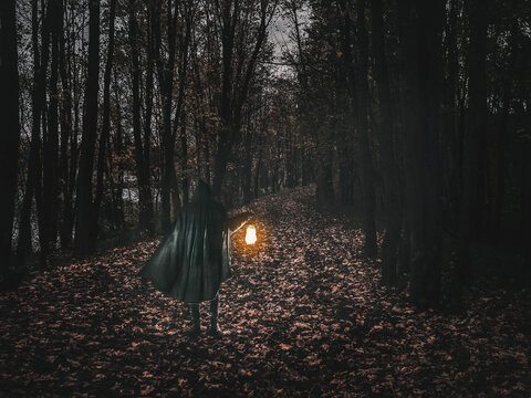 Man in motion in black brilliant hooded cloak holding the glowing lantern on the dark forest