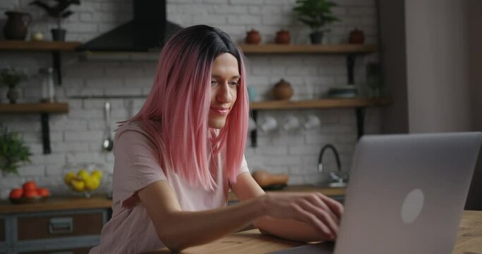 Attractive young transwoman with pink wig types message on laptop and smiles sitting at table in decorated kitchen at home